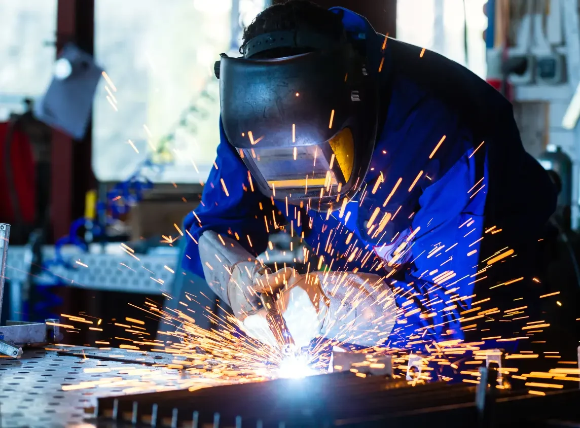A professional welder working in a dark industrial setting, creating bright blue and orange sparks while delivering high-end Customized Fabrication Solutions​ for heavy-duty metal structures.